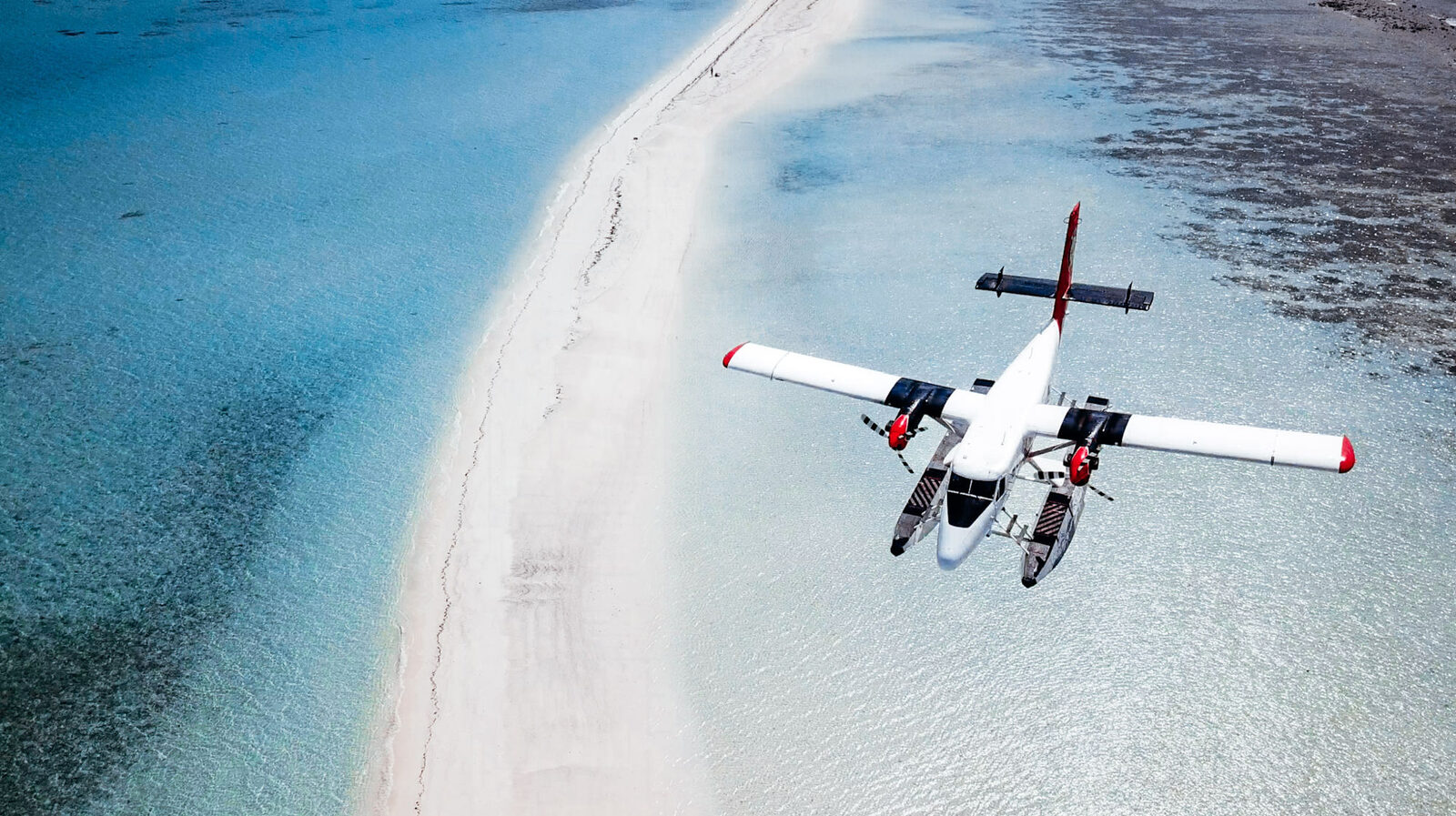 A Trans Maldivian Airways seaplane flying low over a sandbank and shallow reef
